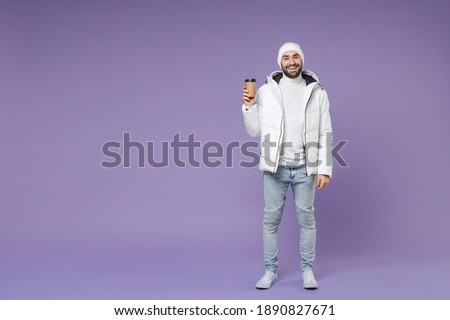Full length of smiling attractive man in warm white windbreaker jacket hat hold paper cup of coffee or tea isolated on purple background studio portrait. People lifestyle cold winter season concept