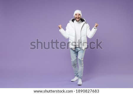 Full length joyful happy man in white windbreaker jacket hat doing winner gesture clenching fist celebrating isolated on purple background studio portrait. People lifestyle cold winter season concept