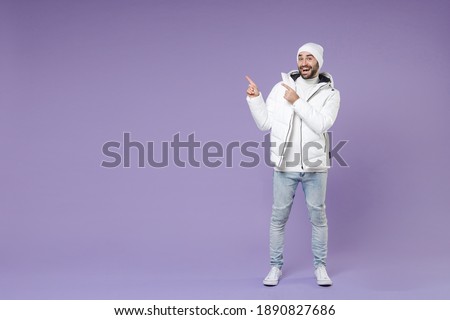 Full length of excited man in warm white padded windbreaker jacket hat pointing index fingers aside isolated on purple color background studio portrait. People lifestyle cold winter season concept