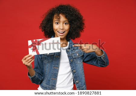 Excited little african american kid girl 12-13 years old in denim jacket point index finger on gift certificate isolated on bright red background children studio portrait. Childhood lifestyle concept