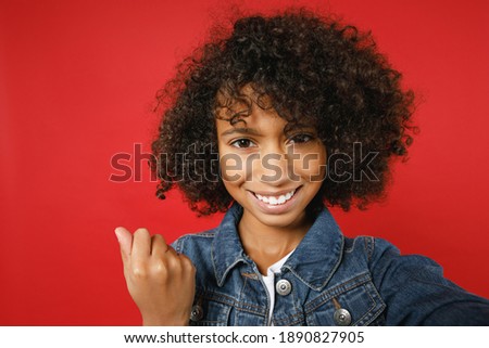Close up of joyful little african american girl 12-13 in denim jacket doing selfie shot on mobile phone doing winner gesture isolated on red background children portrait. Childhood lifestyle concept