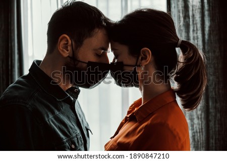 Couple in love with face masks standing next to window and looking at each other. Love during corona virus.