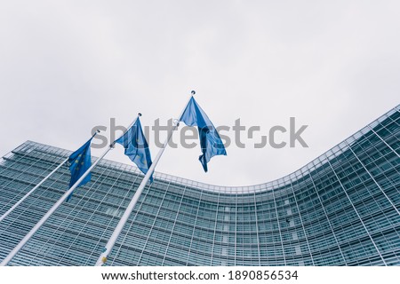 The queue of steeples with blue flags of the European Union against the background of the European Commission building in Brussels, Belgium. EU flag, symbol Royalty-Free Stock Photo #1890856534
