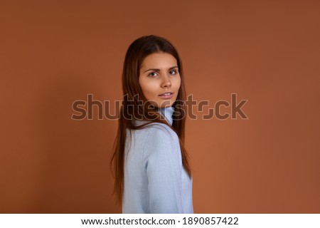 Pretty caucasian woman with dark long hair wearing light blue sweater stands sideways to camera, smiling gently, isolated over brown background