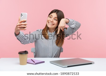 Cute positive woman blogger in striped shirt pointing finger down recording video for followers, asking to subscribe and turn on notifications. Indoor studio shot isolated on pink background