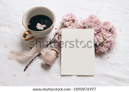 Wedding stationery mock-up. Blank greeting card and cup of coffee. White linen tablecloth background. Pink blossoming cherry tree branches and ribbon. Feminine still life composition. Flat lay,top.