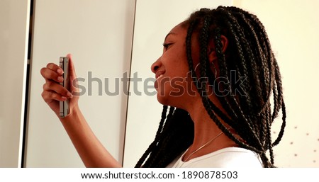 
Teenage adolescent girl taking selfie photo in front of mirror reflection. Black African ethnicity