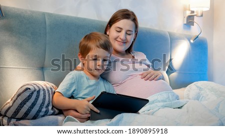 Family lying in bed and watching cartoons on tablet computer at night. Little boy and pregnant mother using digital tablet in bed at night.