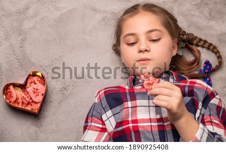 Beautiful girl with pigtails eating cake shape of heart on a grey carpet, romantic valentine day concept, symbol of day Valentine. Copy space. Flat lay