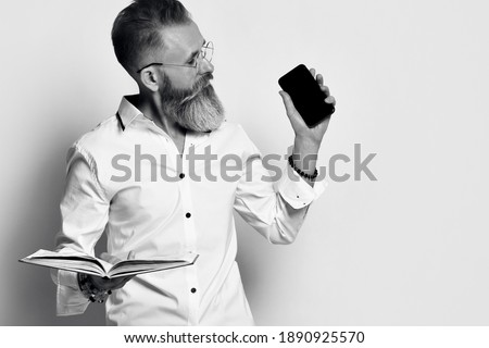 stylish brutal man with a gray beard holds a book or diary in his hands and shows the screen of his mobile phone. black and white photo