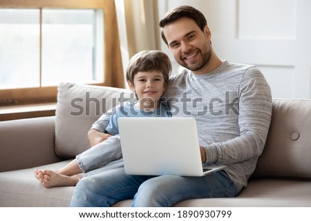 Family portrait of happy young Caucasian dad and little son relax on couch at home using computer. Smiling father and small boy child rest on sofa in living room browse internet on laptop together.