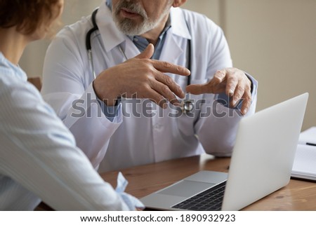 Close up older mature male doctor physician in white uniform gesturing, explaining medical insurance program presentation on computer to focused patient or giving healthcare advice at appointment. Royalty-Free Stock Photo #1890932923