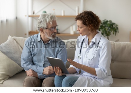 Skilled professional physician doctor prescribing illness treatment, showing health test results on digital computer tablet to concentrated older retired patient at home, sitting together on sofa. Royalty-Free Stock Photo #1890933760