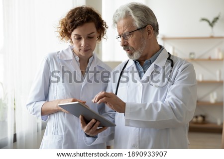 Focused two different generations doctors looking at digital tablet screen, discussing patient's illnesses or diagnosis in clinic. Young nurse assisting older male general practitioner in hospital. Royalty-Free Stock Photo #1890933907