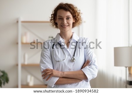 Portrait of smiling young doctor physician in white coat and stethoscope standing in clinic office with folded arms. Happy trusted female therapist looking at camera, waiting for patient in hospital. Royalty-Free Stock Photo #1890934582