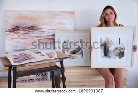 Modern art and lifestyle. Female artist showing her work in the studio. Portrait of young adult artist in her 20s smiling, with her abstract and colorful framed paintings, canvas and drawings.