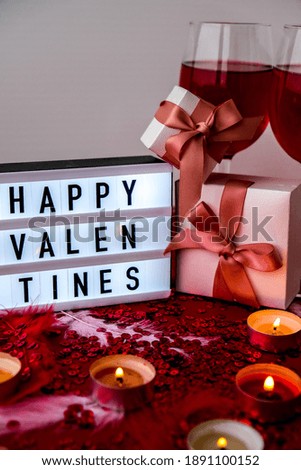 Lightbox with text HAPPY VALENTINES two glasses of red wine and gift boxes and Burning red candles on Red shiny confetti sequinned background with feathers. Saint Valentines Day and Christmas backdrop