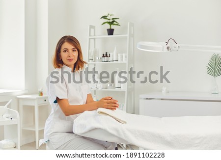 Young positive woman cosmetologist or dermatologist sitting near empty beauticians coach awaiting for client visit and looking at camera in beauty spa salon. Cosmetology and skincare concept Royalty-Free Stock Photo #1891102228