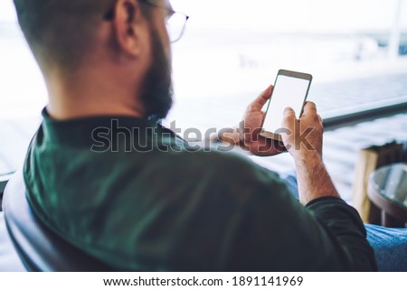 Back view of bearded male in casual clothes sitting in armchair and looking at screen of smartphone while checking social network