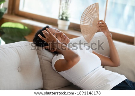 Stressed unhappy young african ethnicity woman lying on comfortable couch, fanning herself. Unhealthy millennial mixed race lady feeling overheated alone at home, summertime hot weather concept. Royalty-Free Stock Photo #1891146178