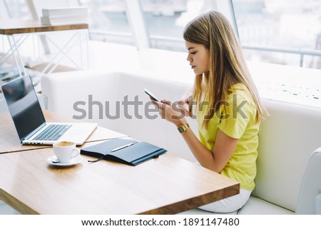Side view of pensive female manager dialing smartphone sitting at table with laptop notepad and cup of coffee in office lounge