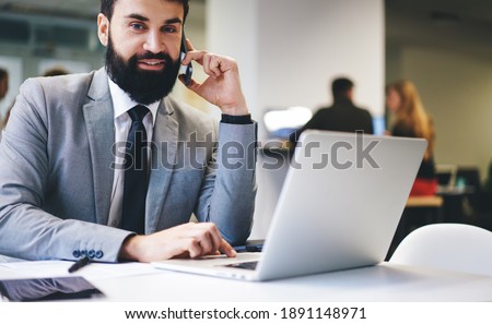 Crop positive entrepreneur in business suit looking at camera sitting at table with laptop while working and having conversation on smartphone during workday