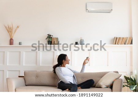 Happy young african american woman turning on air conditioner with remote controller, setting comfortable temperature indoors, using climate control appliance at home, modern domestic tech device. Royalty-Free Stock Photo #1891149022