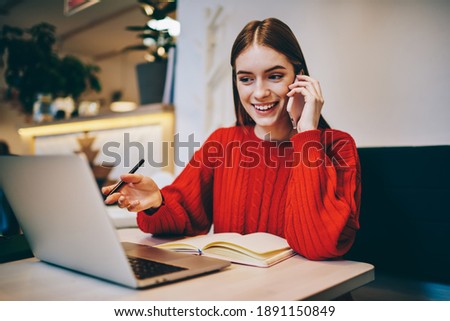 Young female freelancer with toothy smile in red sweater working on laptop and taking notes while using smartphone for calling in cafe