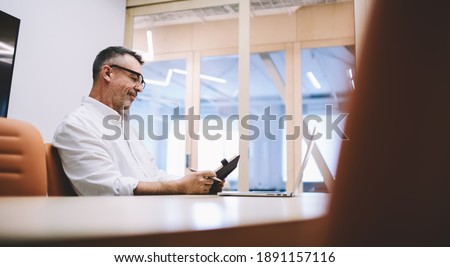Low angle side view of adult businessman using laptop and notebook with pen while sitting at table in modern light workspace Royalty-Free Stock Photo #1891157116