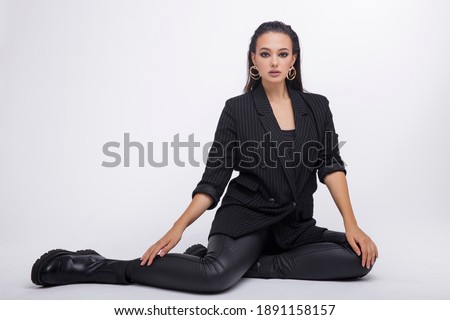Fashion photo of a beautiful elegant young woman in a pretty black jacket, leather pants, boots, posing over white background. The hair is gathered back, dark brunette. Studio Shot