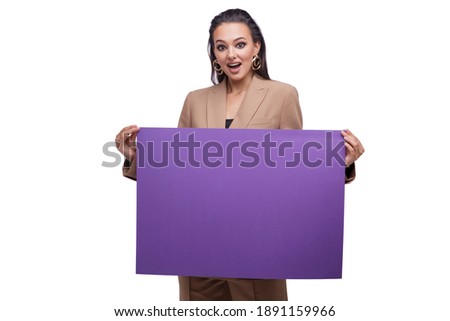 Fashion photo of a beautiful elegant young woman in a pretty oversize brown and beige suit, jacket, pants, posing over white background. Brunette. Showing blank purple big paper.