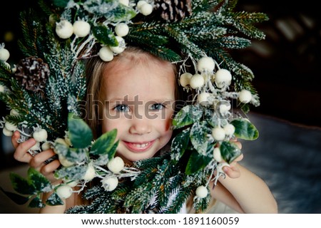 Happy little blonde girl  trying Christmas Wreath. Christmas tree on thge background.