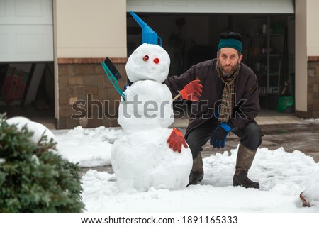 man crouched next to a snowman figure of three large snow balls in the background of the garage. The snowman has a watering can and a brush

