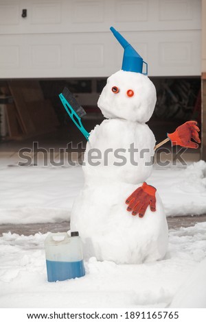 Snowman motorist stands in front of the garage. A watering can, a brush and work gloves are used for decoration. There is a canister with blue antifreeze nearby

