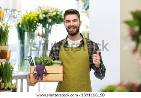 gardening, farming and people concept - happy smiling male gardener in apron with box of garden tools showing thumbs up over flower shop background