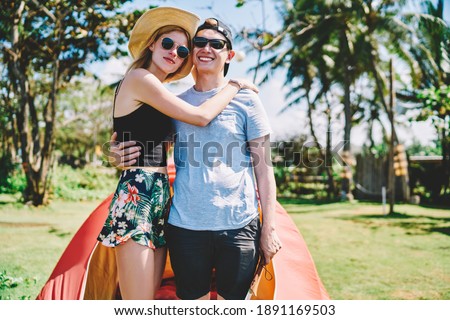 Happy hugging couple of young travellers in summer casual outfit and sunglasses enjoying honeymoon beside camping tent in tropical place