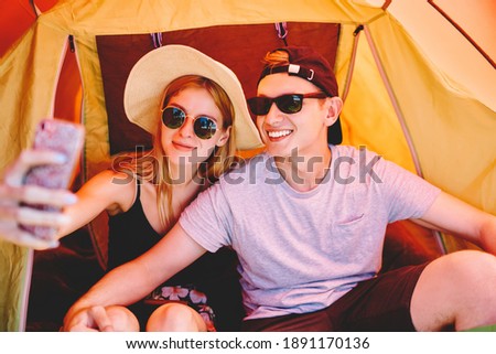 Cheerful tourists in summer casual outfit and sunglasses taking selfie together using mobile phone in camping tent on summer holidays