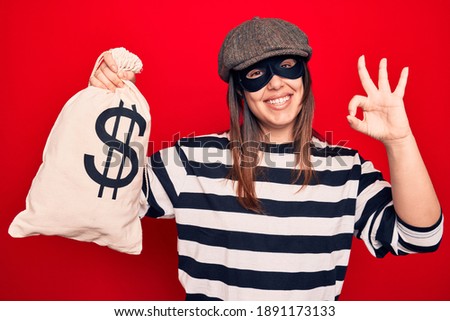 Young beautiful burglar woman wearing cap and mask holding money bag with dollar symbol doing ok sign with fingers, smiling friendly gesturing excellent symbol