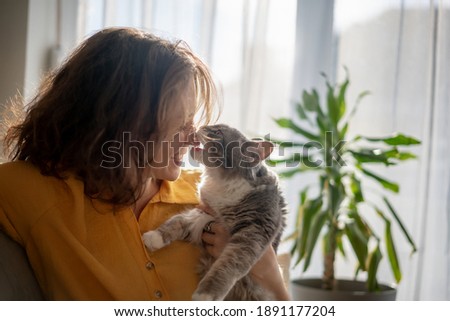Portrait of a young beautiful woman in a yellow shirt hugging kissing with a gray fluffy cat sitting on the sofa Royalty-Free Stock Photo #1891177204