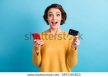 Photo of surprised cute young lady smiling showing credit card telephone prepare pay wear yellow sweater isolated blue color background