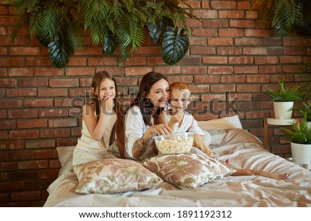 Mom sits on the bed with her son and daughter and watch a movie. A woman, a boy and a girl eat popcorn while watching a movie in the bedroom. The family is resting at home on the weekend