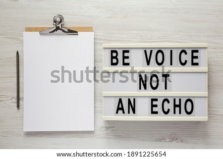 'Be voice not an echo' on a lightbox, clipboard with blank sheet of paper on a white wooden background, top view. Flat lay, overhead, from above. 