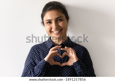 Headshot portrait of smiling young Indian woman isolated on grey studio background show heart love hand gesture or sign. Profile picture of happy ethnic female feel grateful. Support and care concept.
