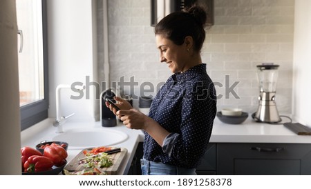 Happy young 20s Indian woman cook healthy vegetarian salad at home kitchen read recipe on cellphone. Smiling ethnic millennial female prepare diet food use smartphone texting or messaging.
