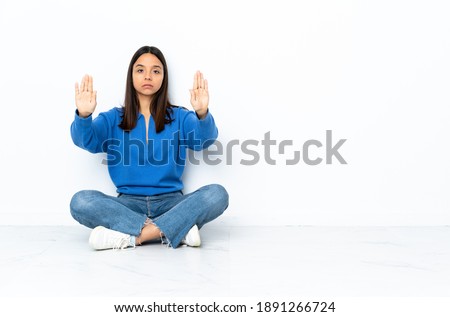Young mixed race woman sitting on the floor isolated on white background making stop gesture and disappointed