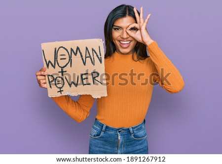 Young latin transsexual transgender woman holding woman power banner smiling happy doing ok sign with hand on eye looking through fingers 