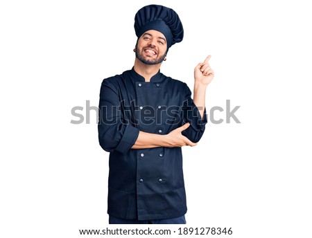 Young hispanic man wearing cooker uniform with a big smile on face, pointing with hand and finger to the side looking at the camera. 