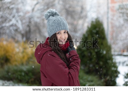 Photo session with a model in the snow, she wears a red coat and a gray hat. It's snowing.

