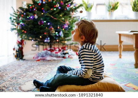 Little adorable preschool kid boy watching television, indoors. Lonely alone child enjoying cartoons on tv during corona virus lockdown quarantine. With Christmas tree on background.