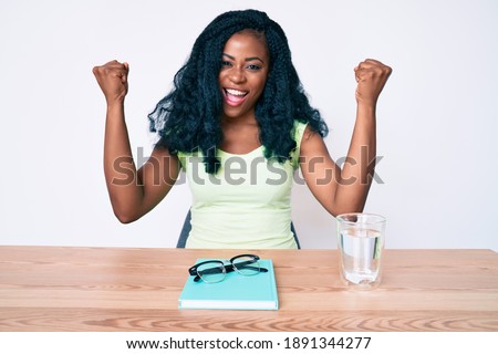 Beautiful african woman sitting on the table stuying for university screaming proud, celebrating victory and success very excited with raised arms 